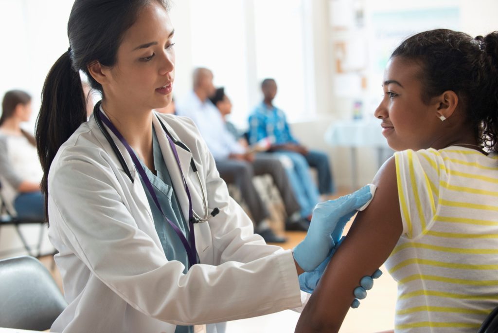 A woman in white coat with a stethoscope around her neck swabs the arm of a teenage girl prior to administering a vaccine