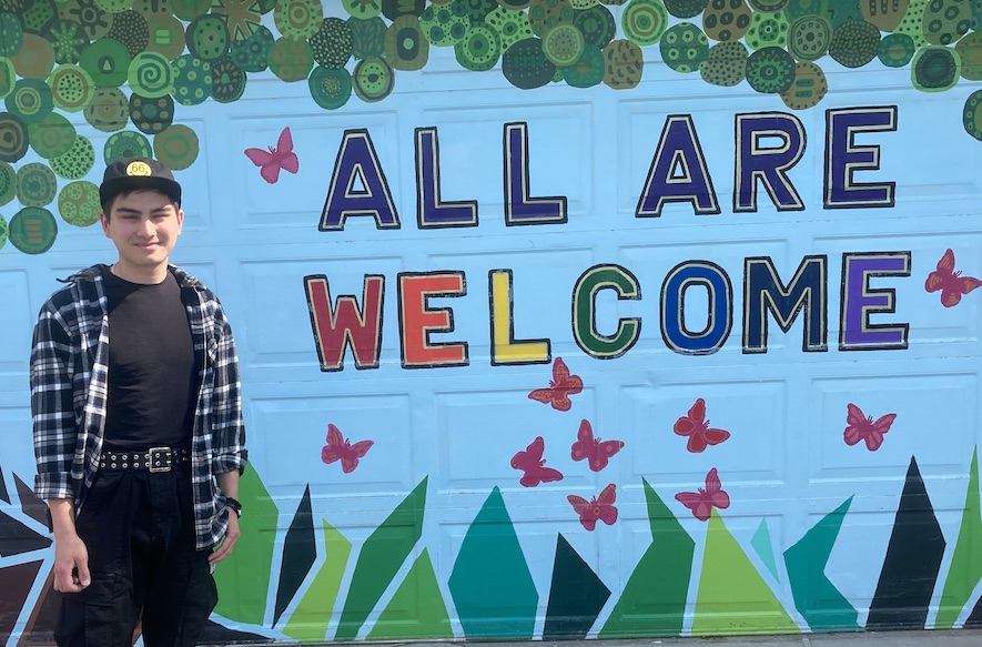 Michael Finch stands in front of a colorful wall at the Eden Prairie Art Center that states: in colorful letters: "All are Welcome" 