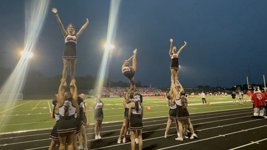 Eden Prairie High School (EPHS) cheerleaders perform on sidelines of football field at a nighttime game.