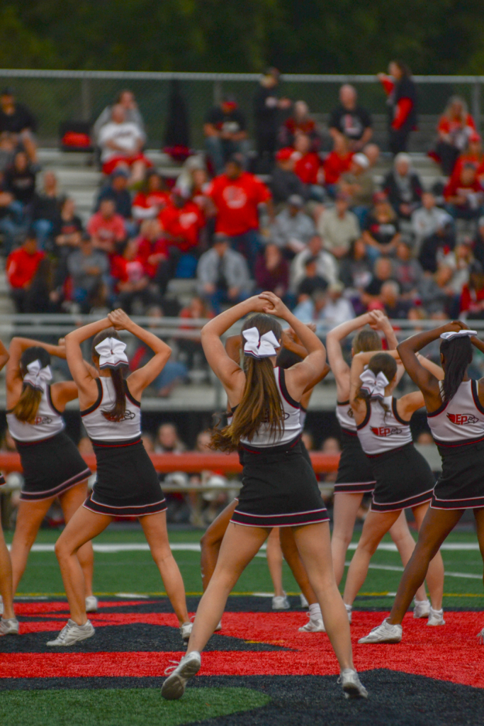 Eden Prairie High School (EPHS) cheerleaders seen from the back as they perform on football field facing people in the bleachers.